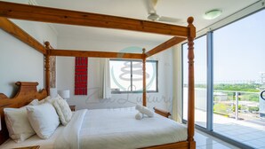 A four-poster bed with a wooden frame, white bedding, and a view of the city through large windows. - Zen Mitchell Rooftop Outdoor Dining Oceanview (Darwin)