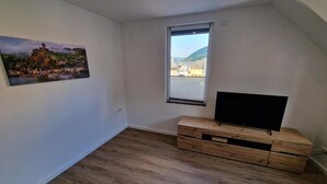 Ein Zimmer mit Holzboden, einem Fenster mit Blick auf eine Hügel- und Stadtlandschaft und einem Flachbildfernseher auf einem Holz-TV-Möbel.