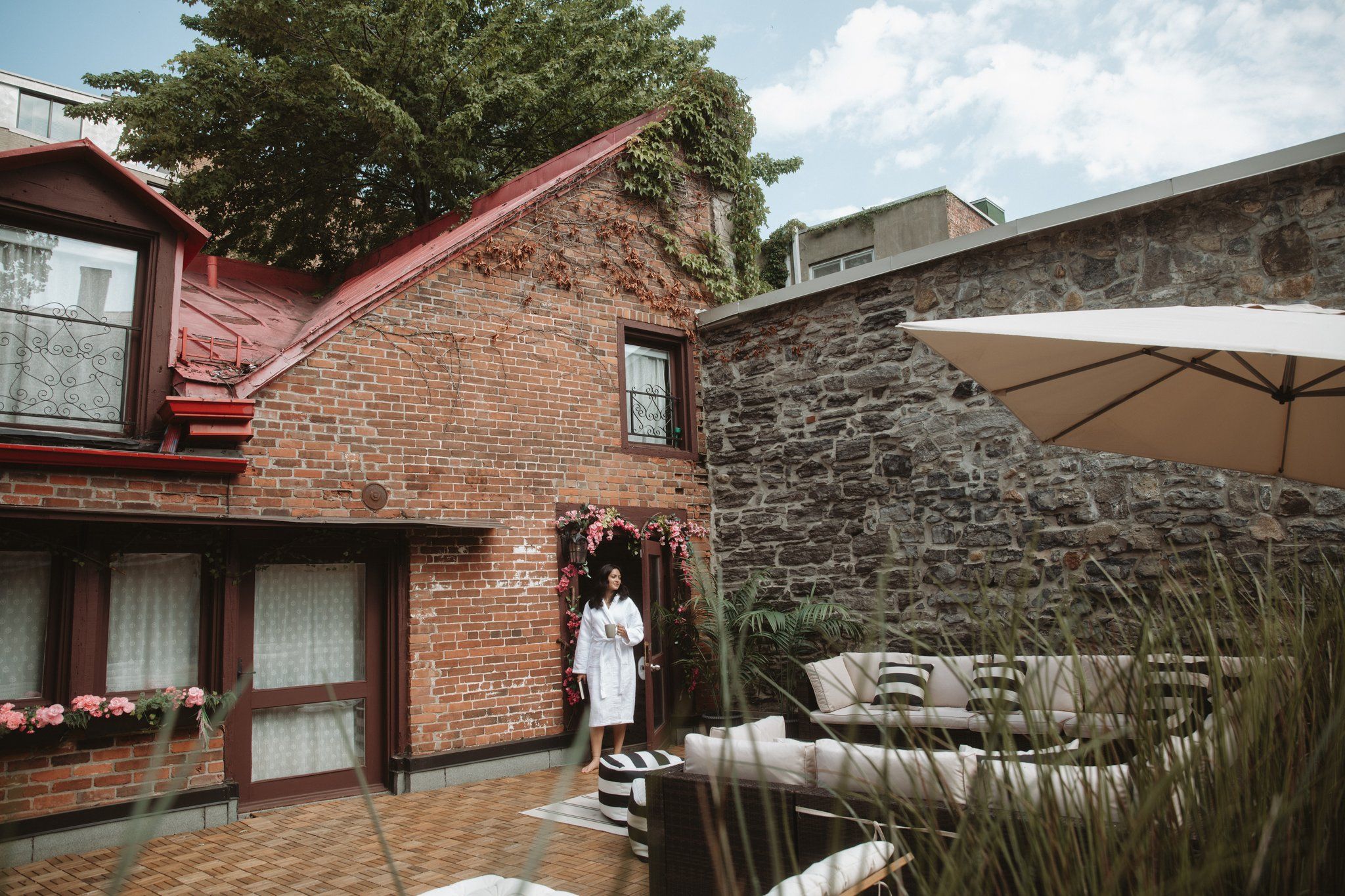 A brick house with a stone wall, a covered patio with outdoor furniture, and a person standing by the entrance.