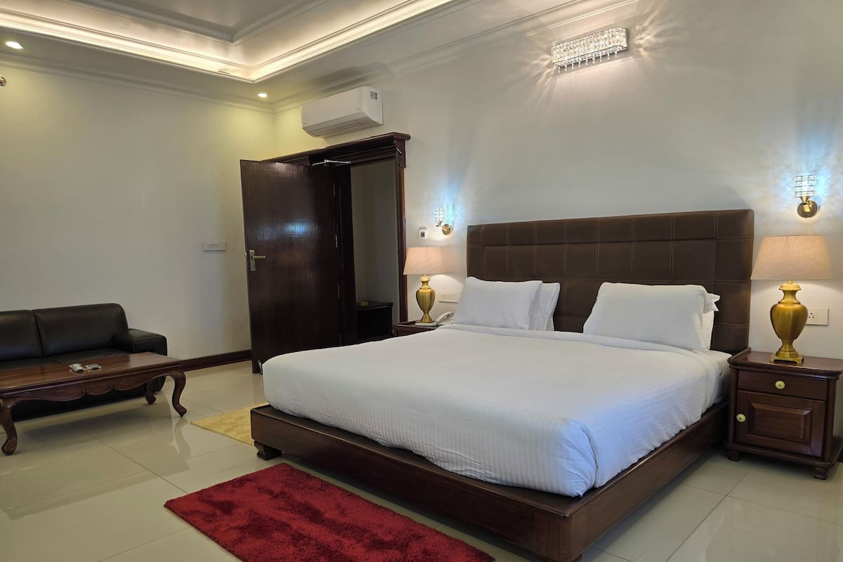 A well-lit bedroom with a large bed, a wooden nightstand, a bedside table, a red rug, a dark wooden door, and a wall-mounted air conditioner.