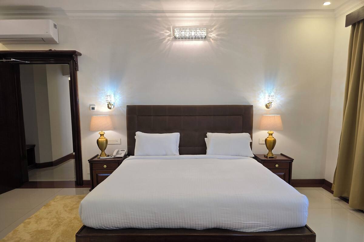 A hotel room with a large bed, two bedside tables with lamps, a wall-mounted air conditioner, and a ceiling light fixture.