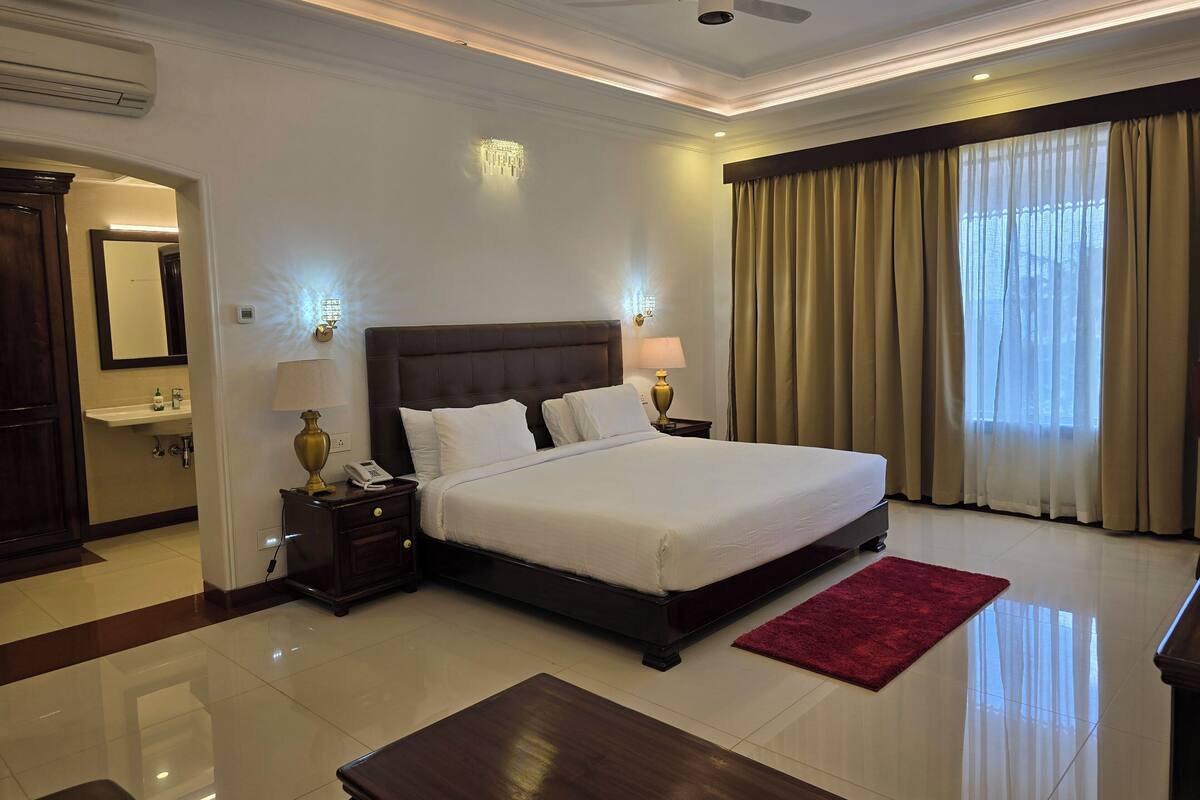 A hotel room with a large bed, bedside lamps, a red rug, a ceiling fan, and a view of the outdoors through the window.