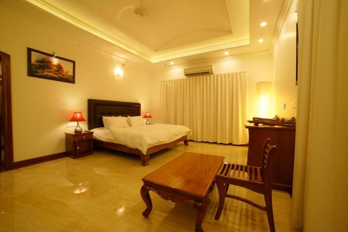 A hotel room with a bed, a wooden table, a chair, a nightstand with a lamp, and a painting on the wall.