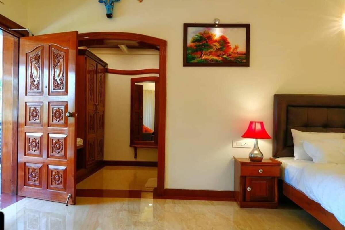A hotel room with a wooden door, a bed with white linens, a nightstand with a red lamp, and a painting on the wall.