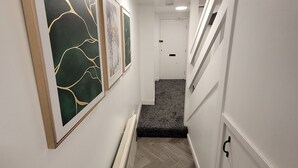 A narrow hallway with white walls, a wooden floor, and a radiator. There are three framed artworks on the left wall.