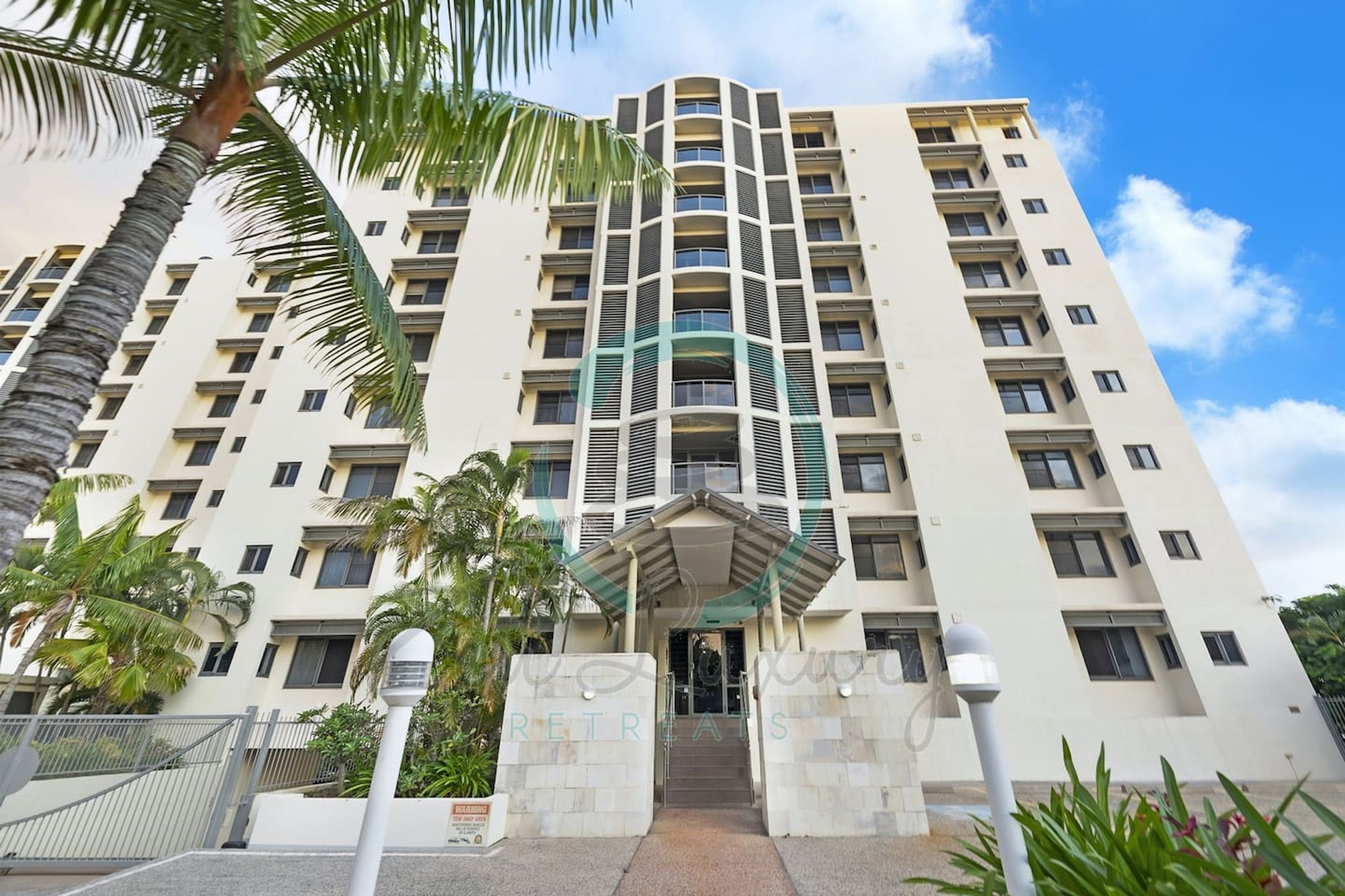 A multi-story hotel with a central entrance, palm trees, and a clear sky.