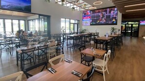 A spacious dining area with multiple tables and chairs, large windows, and a flat-screen TV mounted on the wall. - Zenhouse Stylish Lawn Workspace Outdoordining (Muirhead)