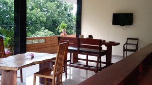 Une pièce spacieuse avec un long banc en bois, une table en bois et des chaises. Un téléviseur est fixé au mur et il y a un balcon agrémenté de plantes.