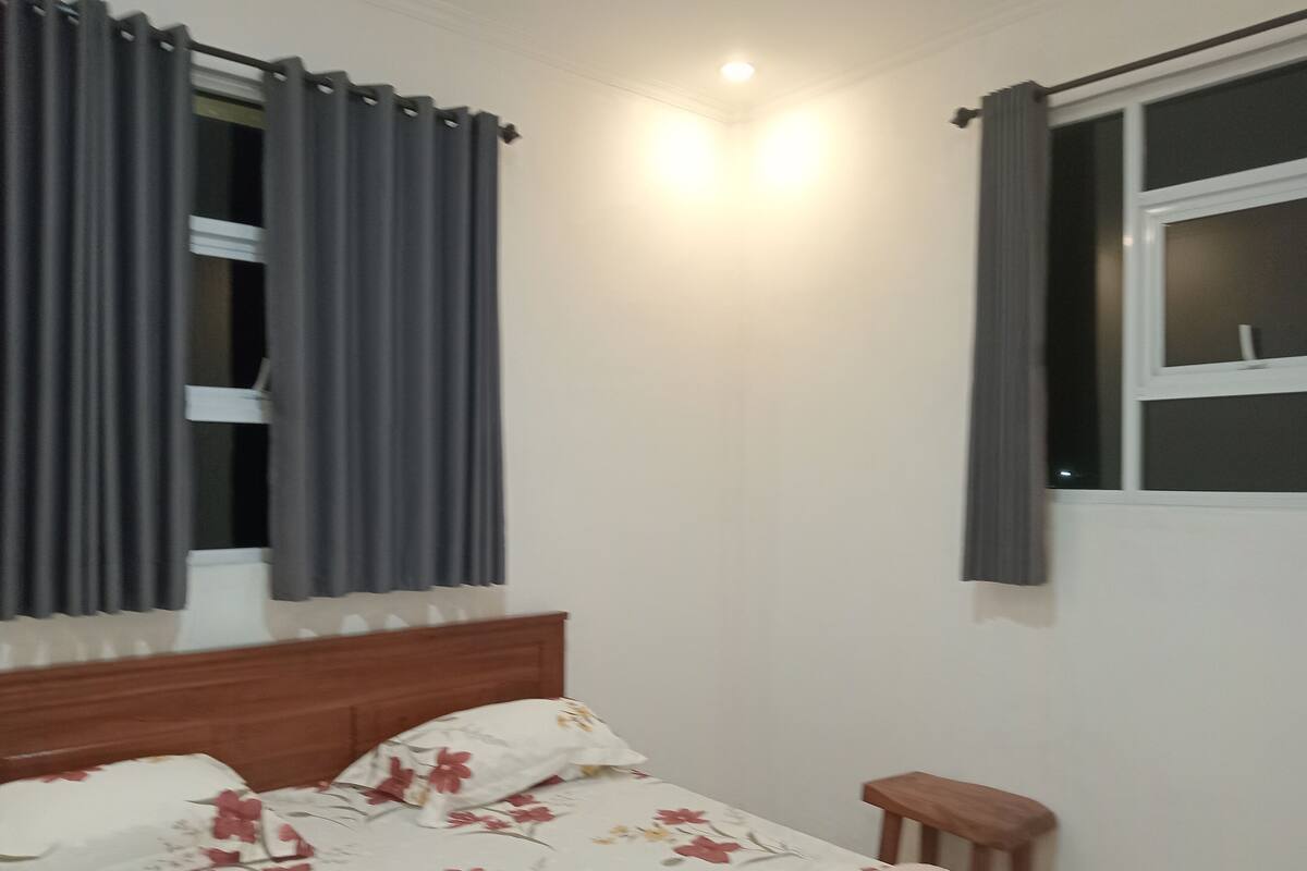 A bedroom with a bed, a nightstand, a window with curtains, and a wall-mounted light fixture.