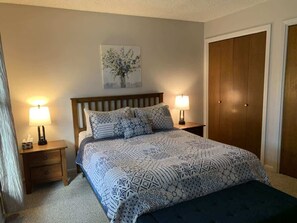A bedroom with a bed, two bedside tables with lamps, a wooden headboard, a wall-mounted artwork, and a wooden wardrobe.