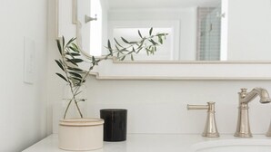 Un baño con encimera blanca, un espejo grande, una planta en maceta, una taza negra y un mueble de madera con cajones.