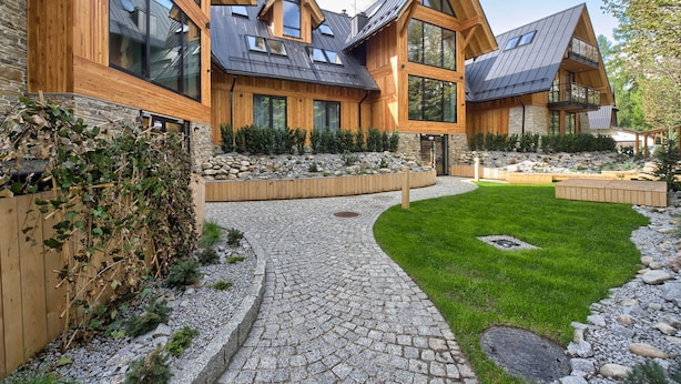 A modern house with a stone facade, wooden accents, and a stone pathway leading to a grassy area.
