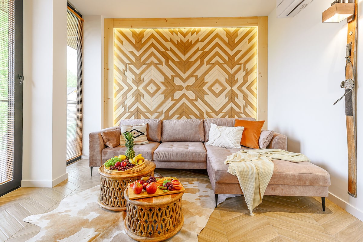 A modern living room with a herringbone-patterned wall, a sectional sofa, and two round wooden side tables with fruit and snacks.