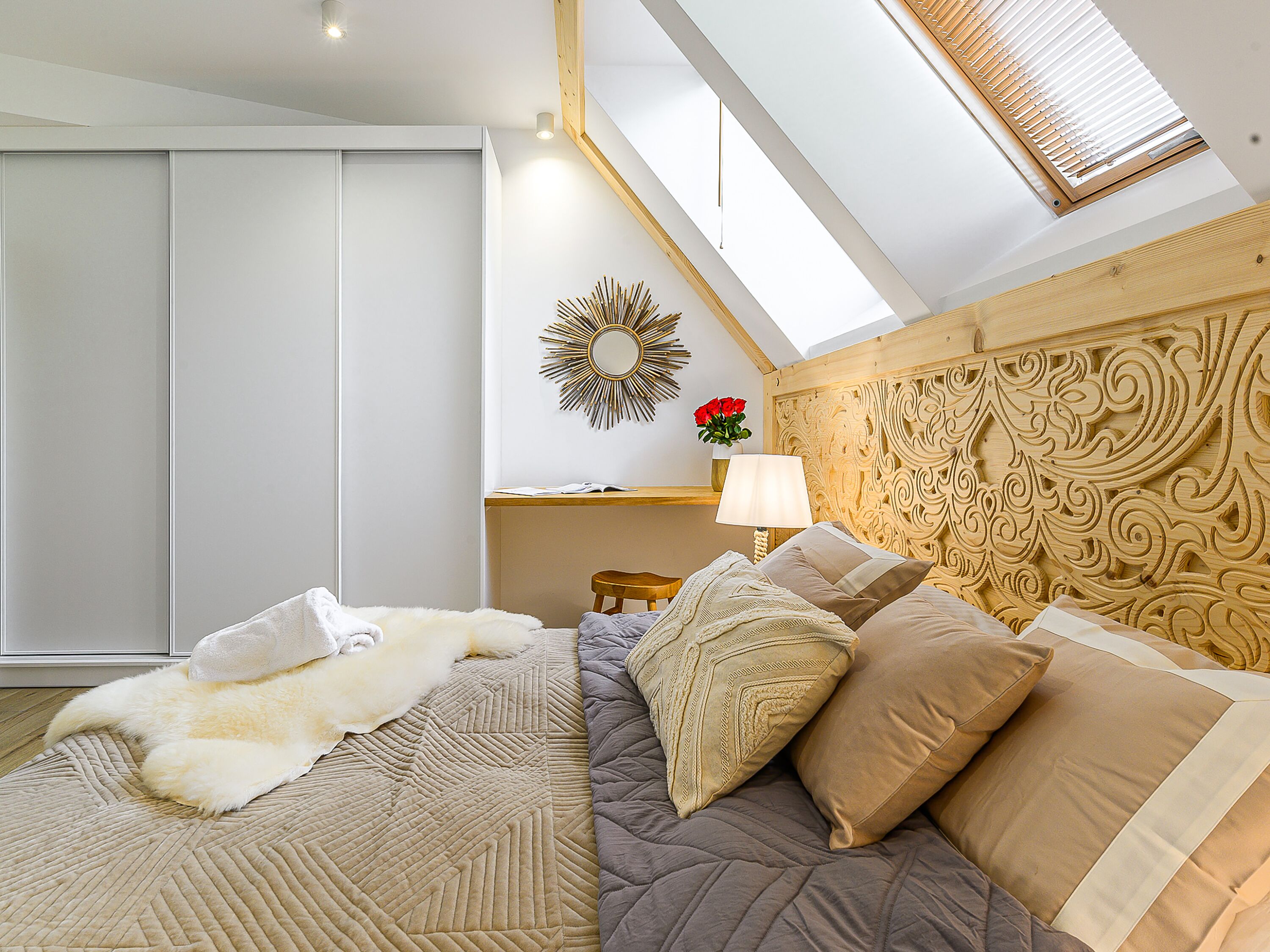 Un dormitorio con una cama, cabecera de madera, mesita de noche con lámpara, espejo y ventana con persianas.