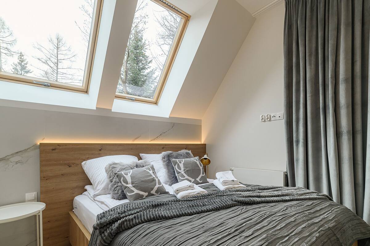 A bedroom with a skylight, a bed with grey bedding, a bedside table, and a curtain.