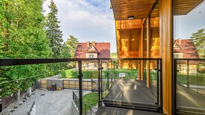 A wooden balcony with a glass railing overlooking a cobblestone path and greenery.