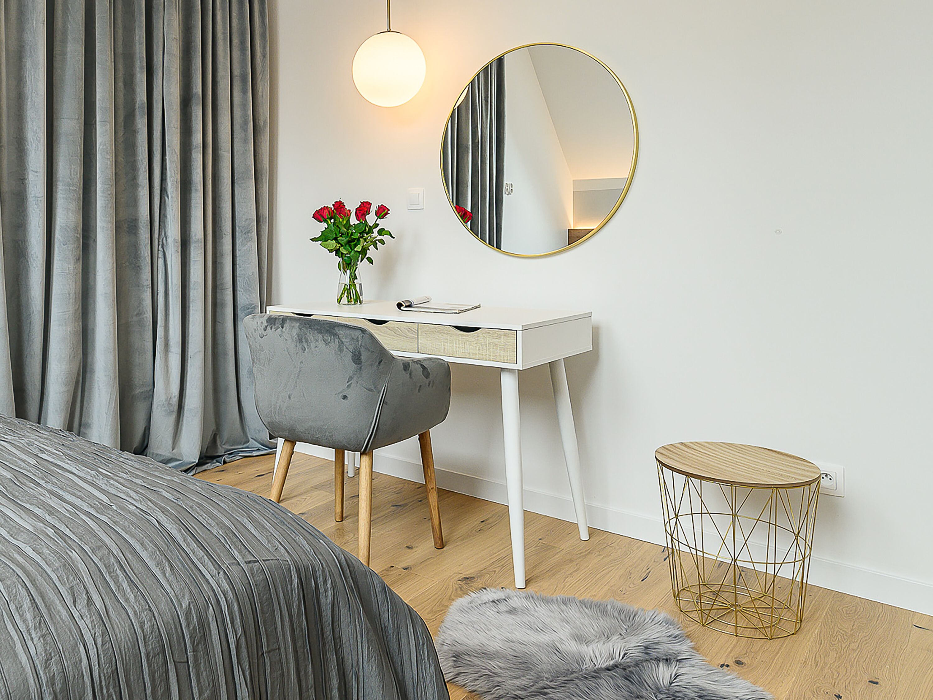 Un dormitorio con cama, escritorio, silla, espejo redondo, pequeña mesa redonda y un florero con flores.
