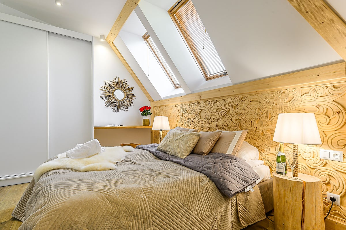 A bedroom with a sloped ceiling, a bed with multiple pillows, a bedside table with a lamp, and a decorative mirror on the wall.