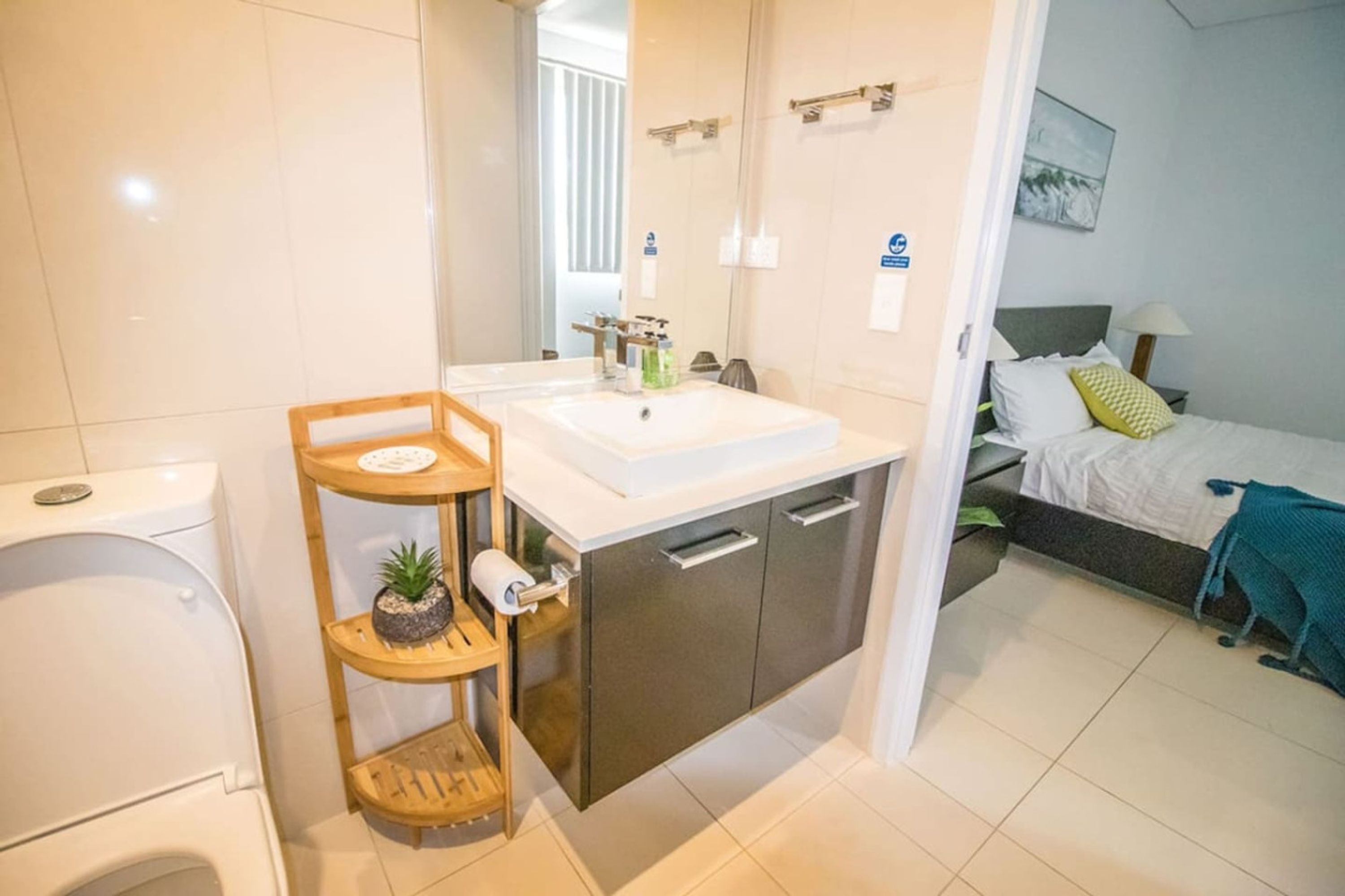 A modern bathroom with a toilet, sink, and shower. There is a wooden shelf with a plant and a towel. The room has a bed with a green blanket and a painting on the wall.