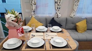 Une table à manger dressée pour quatre personnes, avec de l’argenterie blanche, des couverts et des verres, placée dans une pièce où se trouvent un canapé gris et des rideaux à motifs.