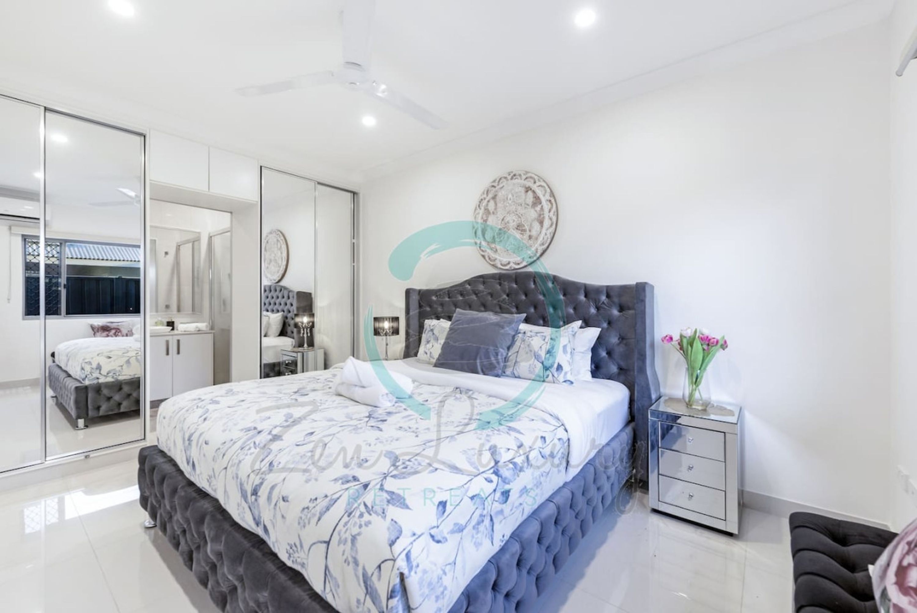 A modern bedroom with a large bed, a wardrobe, a vanity, and a nightstand with a vase of flowers.