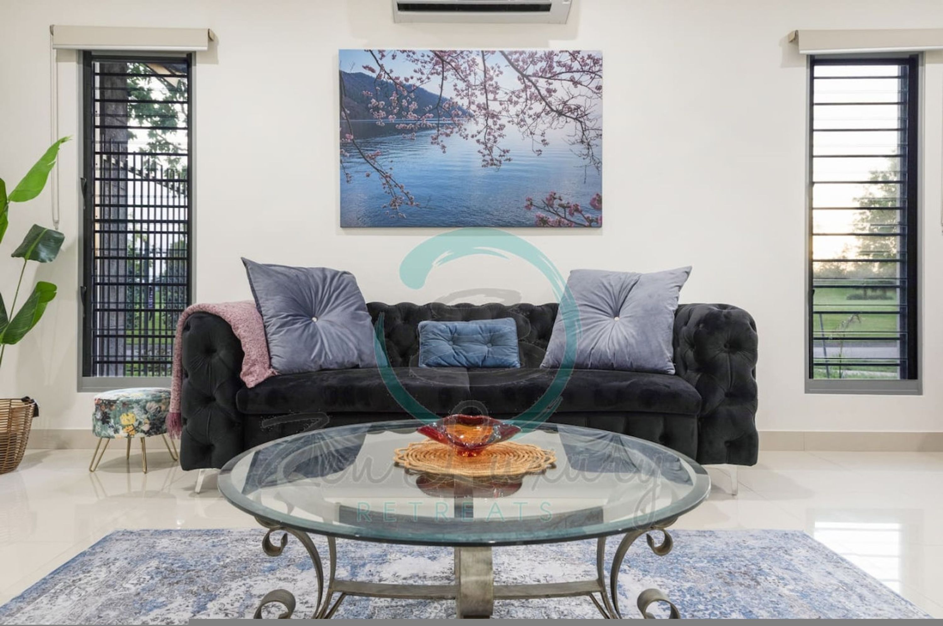 A living room with a black sofa, two cushions, a glass coffee table, and a painting of cherry blossoms.