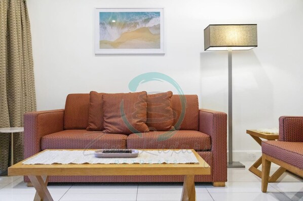 A living room with a patterned sofa, a coffee table, a lamp, and a framed picture on the wall. - Sunny Shores Sea Front City Views Gym Pool (Darwin)