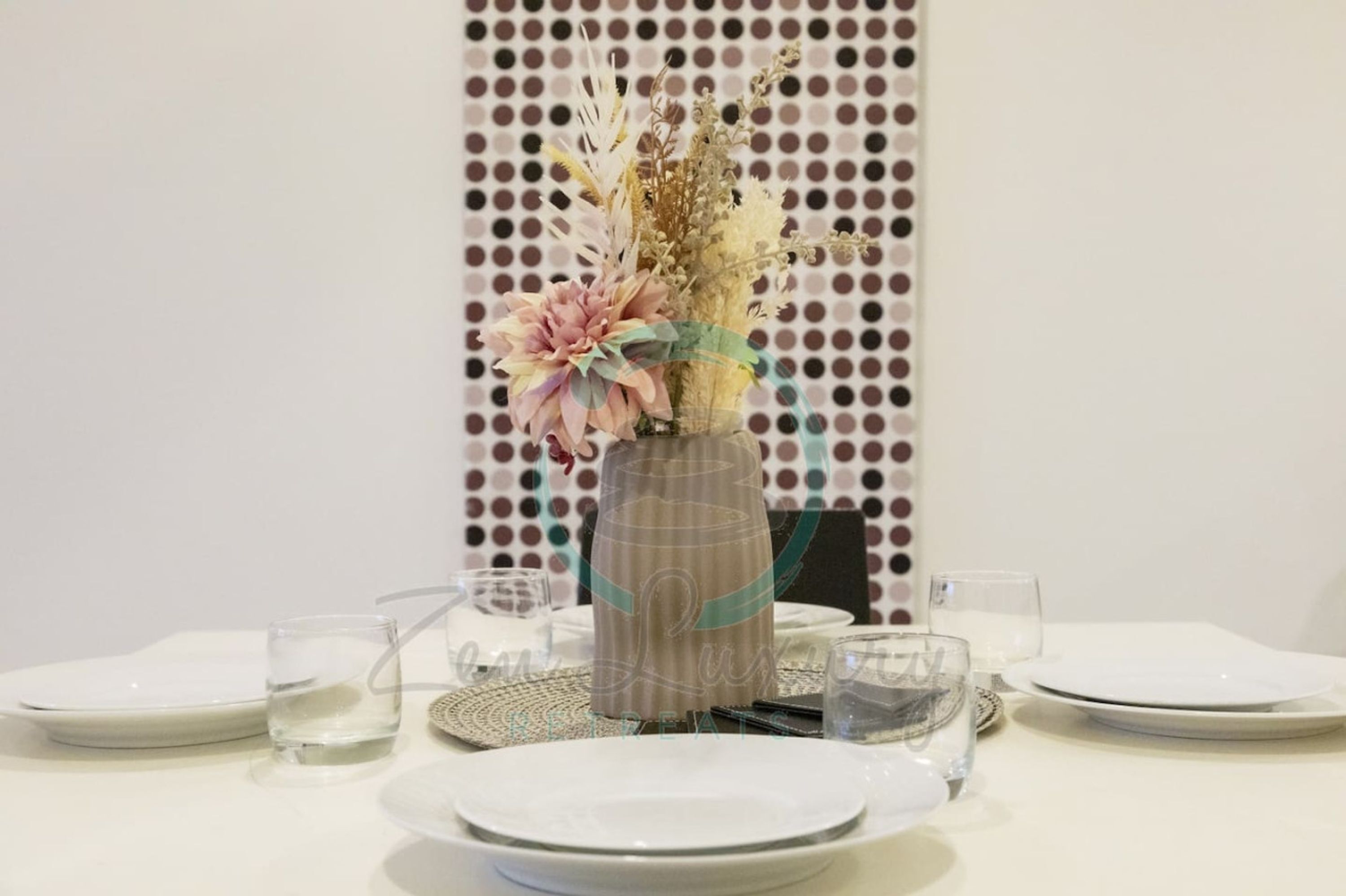 Een eettafel gedekt met witte borden, heldere glazen en een decoratieve vaas met gedroogde bloemen.