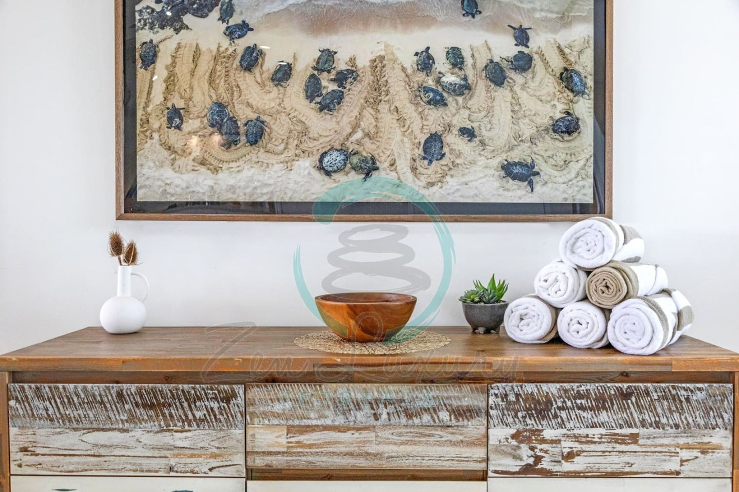 A wooden console table with a rustic finish, a framed artwork depicting prehistoric creatures, a white vase, a small potted plant, and rolled towels.