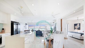 A modern kitchen with white cabinets and a large island, a dining area with a white table and chairs, and a living room with a sofa and a TV. - Zen Skytower Harbourview -exclusive 3br Family Apt (Darwin)