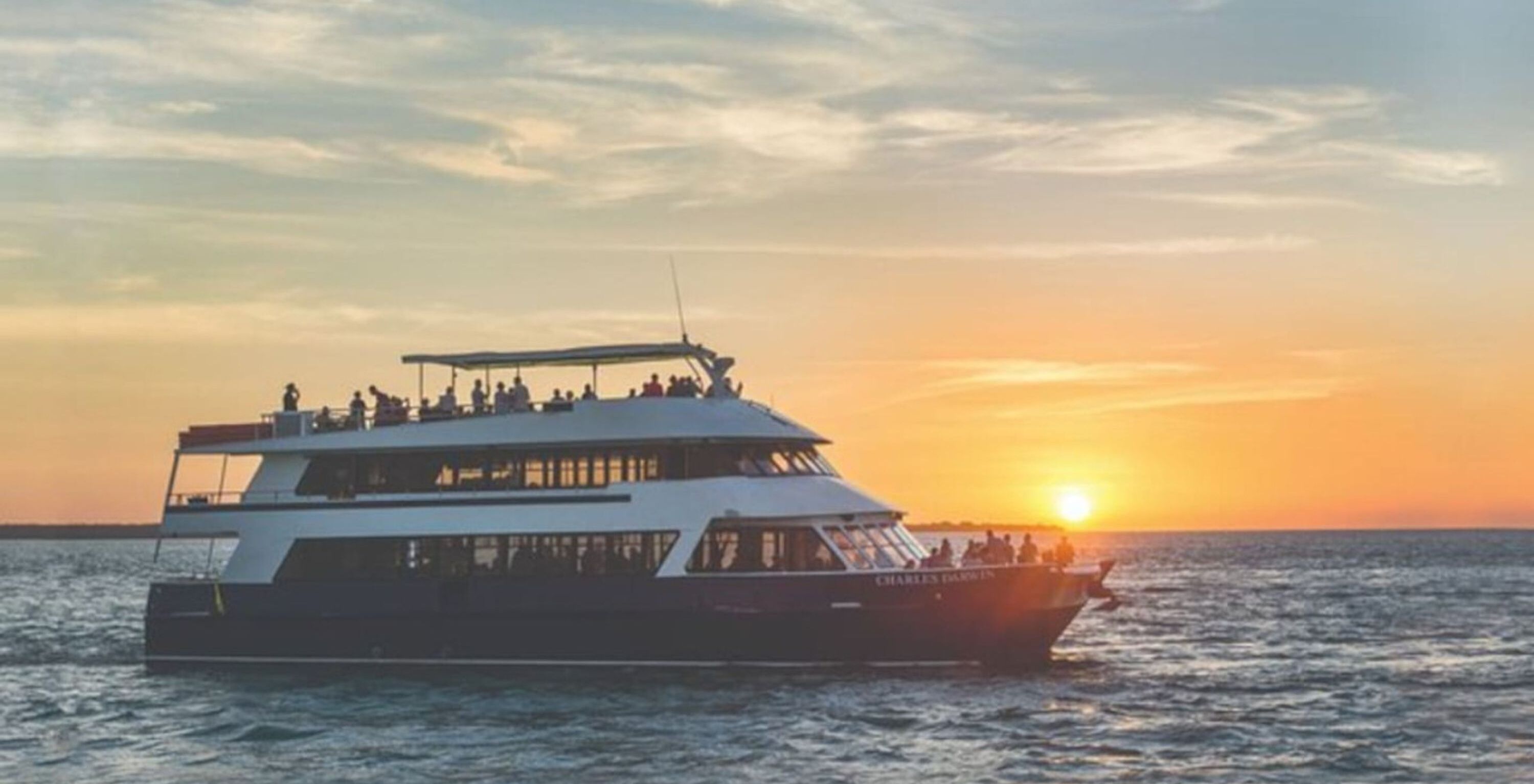 A large, multi-deck boat with the name "Charles Darwin" on the side, sailing on the water during sunset.