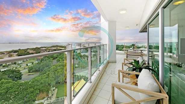 A balcony with a view of a sunset over the ocean, a glass railing, and a seating area with cushions. - Zen Skytower Harbourview -exclusive 3br Family Apt (Darwin)