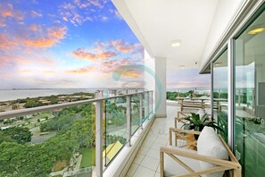 A balcony with a view of a sunset over the ocean, a glass railing, and a seating area with cushions. - Zen Skytower Harbourview -exclusive 3br Family Apt (Darwin)
