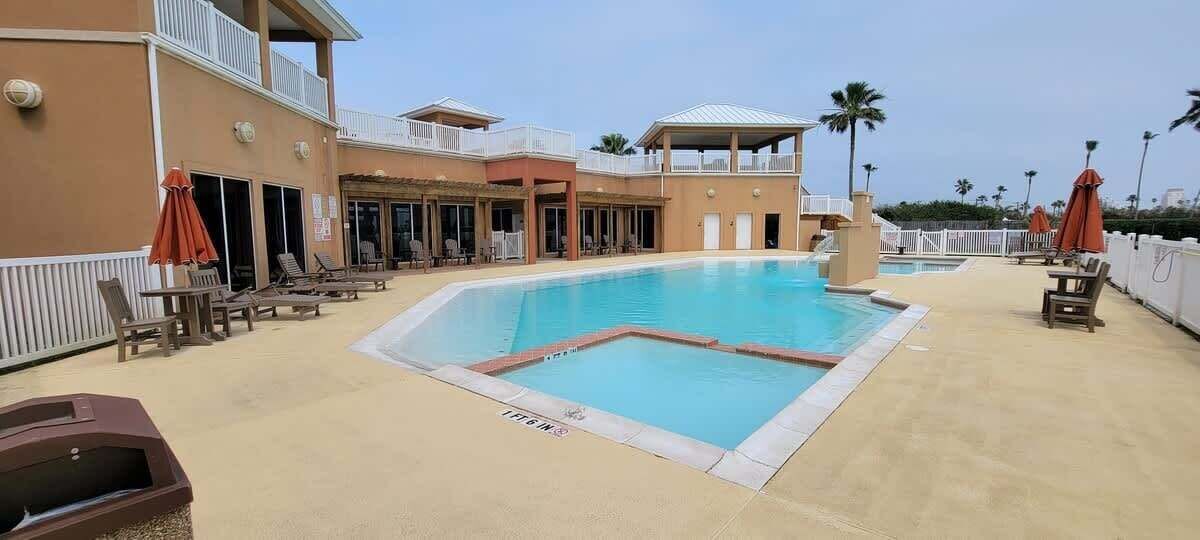 Une piscine entourée de chaises longues et de parasols, le tout bordé d’un bâtiment doté de balcons et d’un espace couvert.