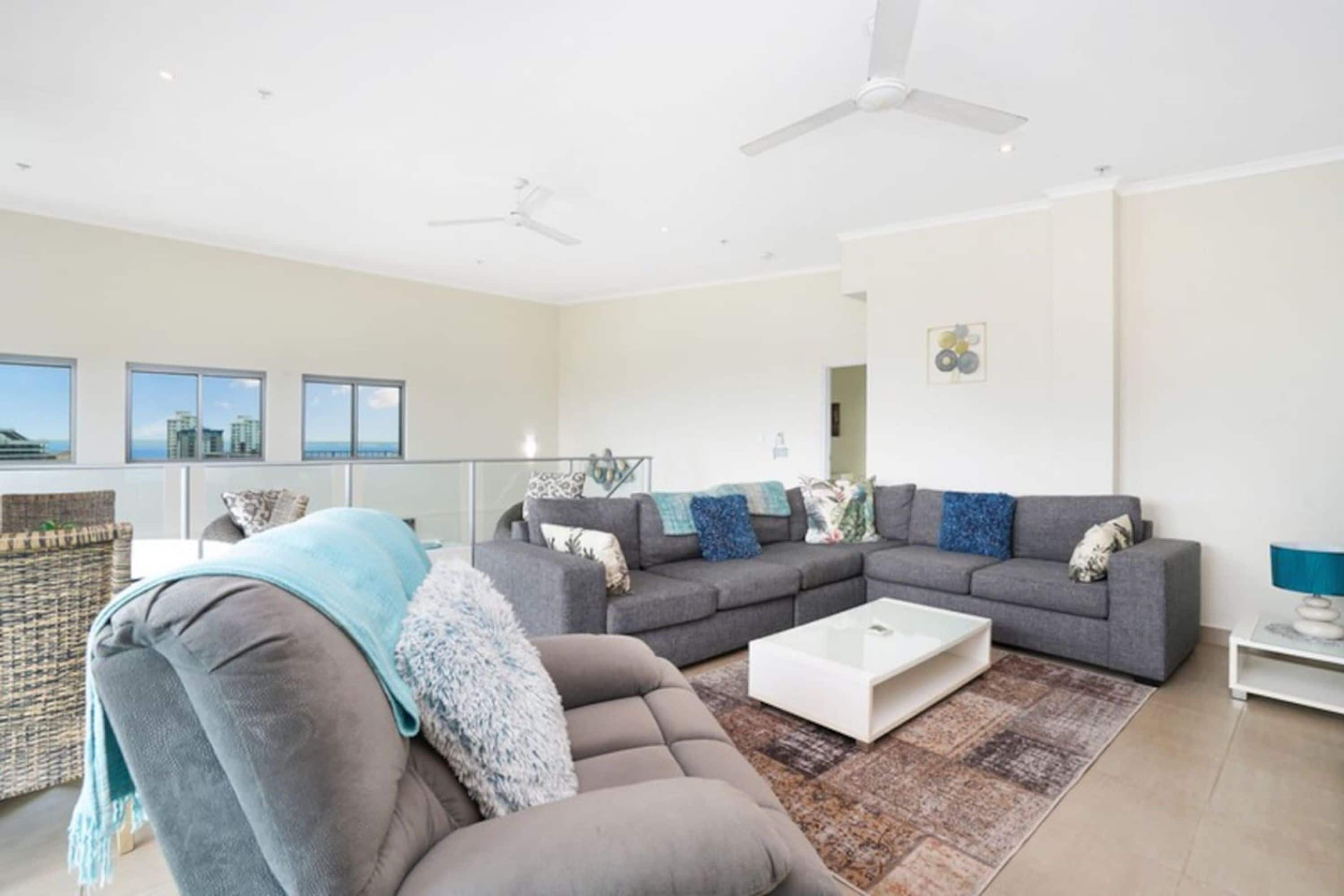 A modern living room with a grey sectional sofa, a white coffee table, and a patterned rug. There are large windows offering a view of the city skyline.
