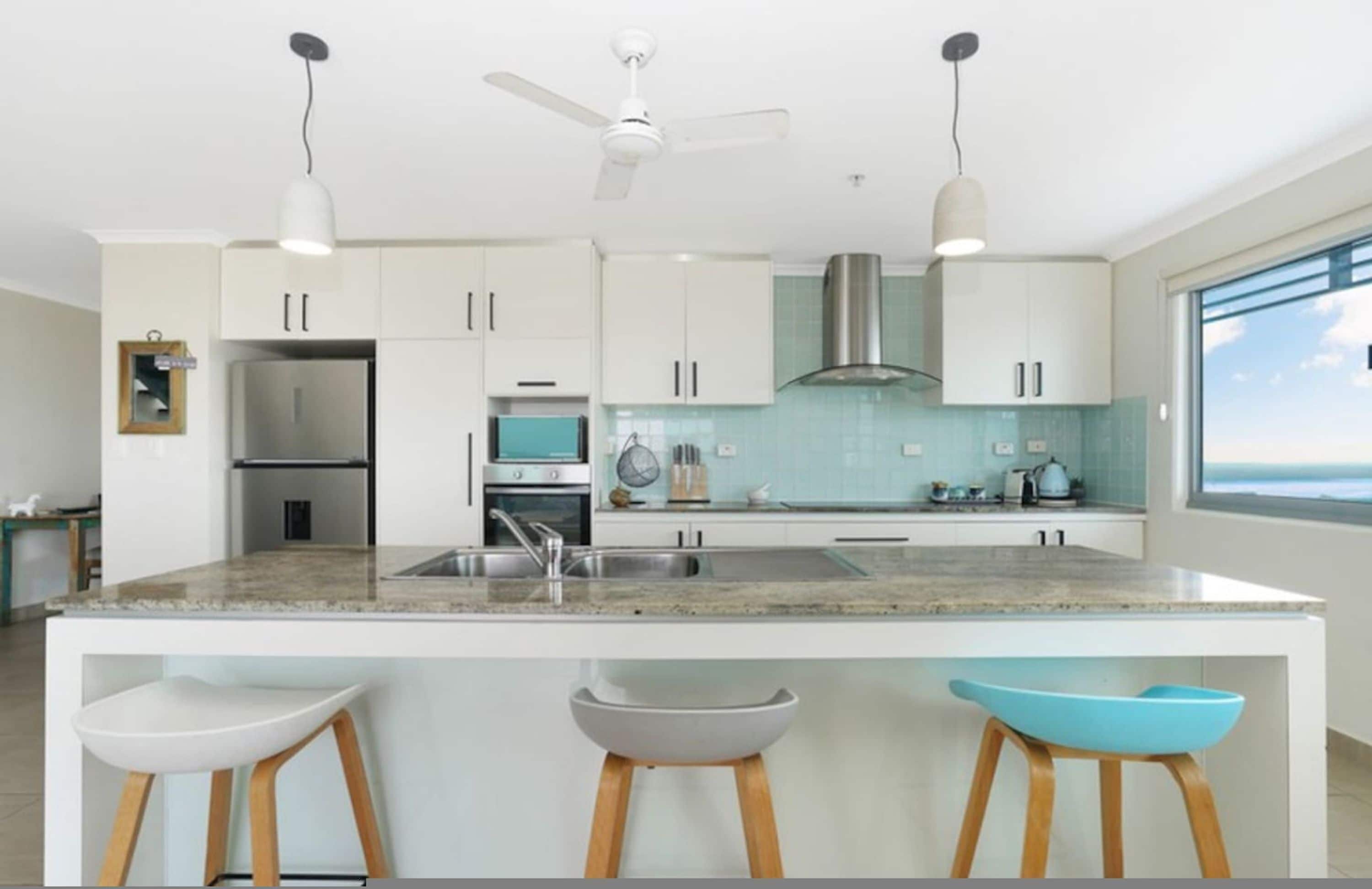 A modern kitchen with white cabinets, a large island with bar stools, stainless steel appliances, and a view to the outside.