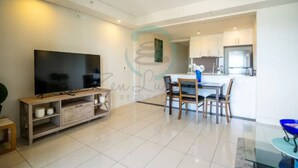 A modern living room with a flat-screen TV, a dining area with a table and chairs, and a kitchen in the background. - Zen Ocean Pebble 1-br 1-ba Sea View Retreat (Darwin)