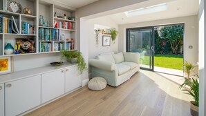 A modern living room with a white sofa, a bookshelf filled with books, and a view of a garden through a sliding glass door. - The Beach House - sleeps 8 guests  in 4 bedrooms (Bembridge)