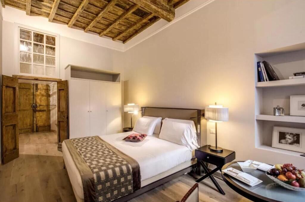 A bedroom with a wooden ceiling, a bed with white linens, a bedside table with a lamp, and a shelf with books and a framed picture.