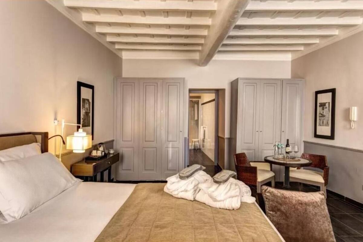 A hotel room with a bed, a desk with a lamp, a chair, a small table with a bottle and glasses, and a wardrobe.