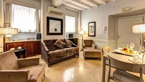 A spacious living room with a sofa, armchairs, a dining table set for two, and a kitchen area in the background.
