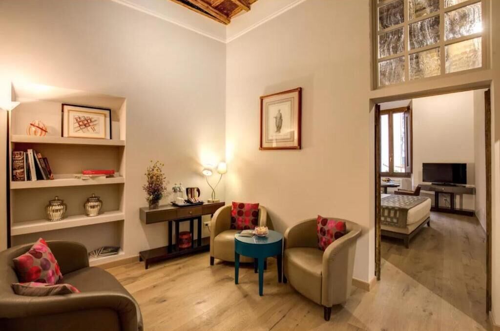 A modern living room with a sofa, armchairs, a coffee table, and a TV. There are shelves with books and decorative items.