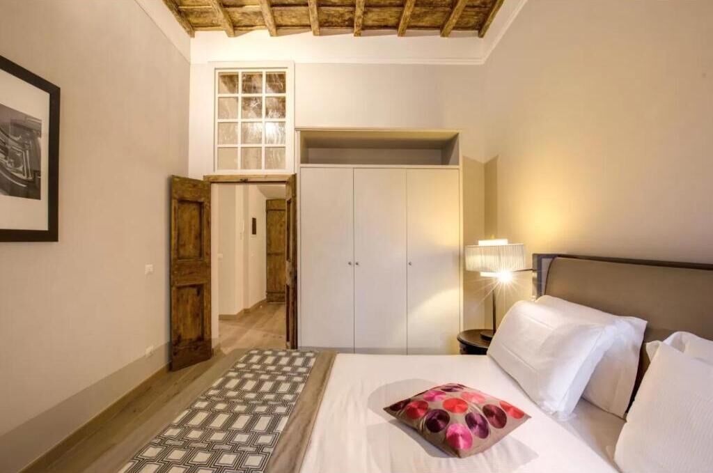 A bedroom with a bed, a nightstand, a lamp, a wardrobe, and a door leading to another room.