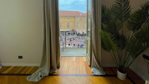 A room with a wooden floor, a large potted plant, and a view of a building with a red roof through open curtains. - Best View Boutique Apartments  (Bergamo)
