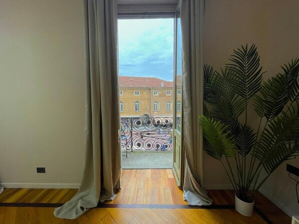 Una habitación con piso de madera, una planta grande en maceta y vista a un edificio con tejado rojo a través de cortinas abiertas.