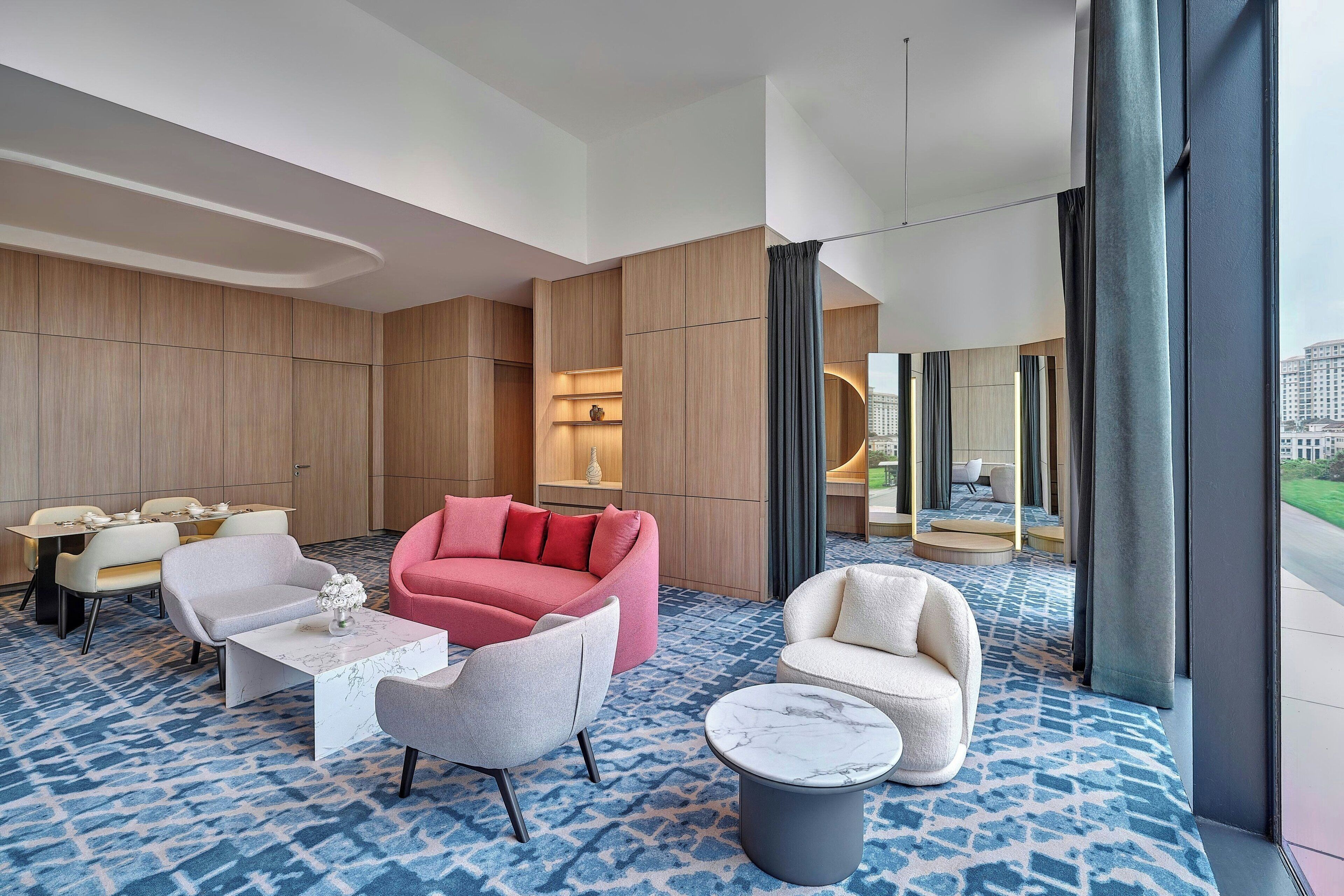 Une chambre d’hôtel moderne avec un tapis bleu à motifs, un canapé rose, un fauteuil blanc, une table basse en marbre et un coin repas avec des chaises et une table blanches.
