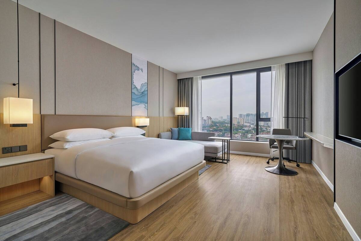 Une chambre d’hôtel moderne dotée d’un grand lit, de tables de chevet, d’un bureau avec une chaise et d’une grande fenêtre donnant sur la ville.