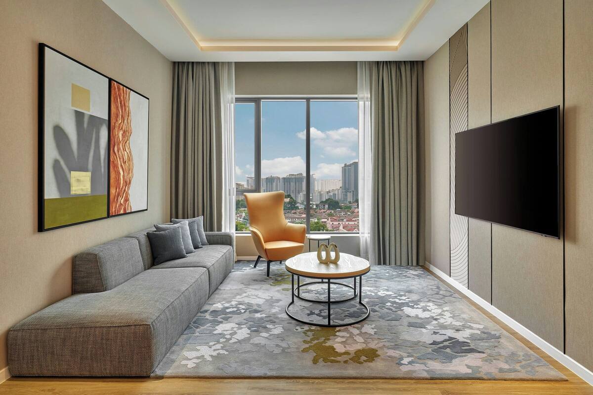 Une chambre d’hôtel moderne dotée d’une grande fenêtre, d’un canapé, d’un fauteuil, d’une table basse ronde et d’un téléviseur à écran plat.