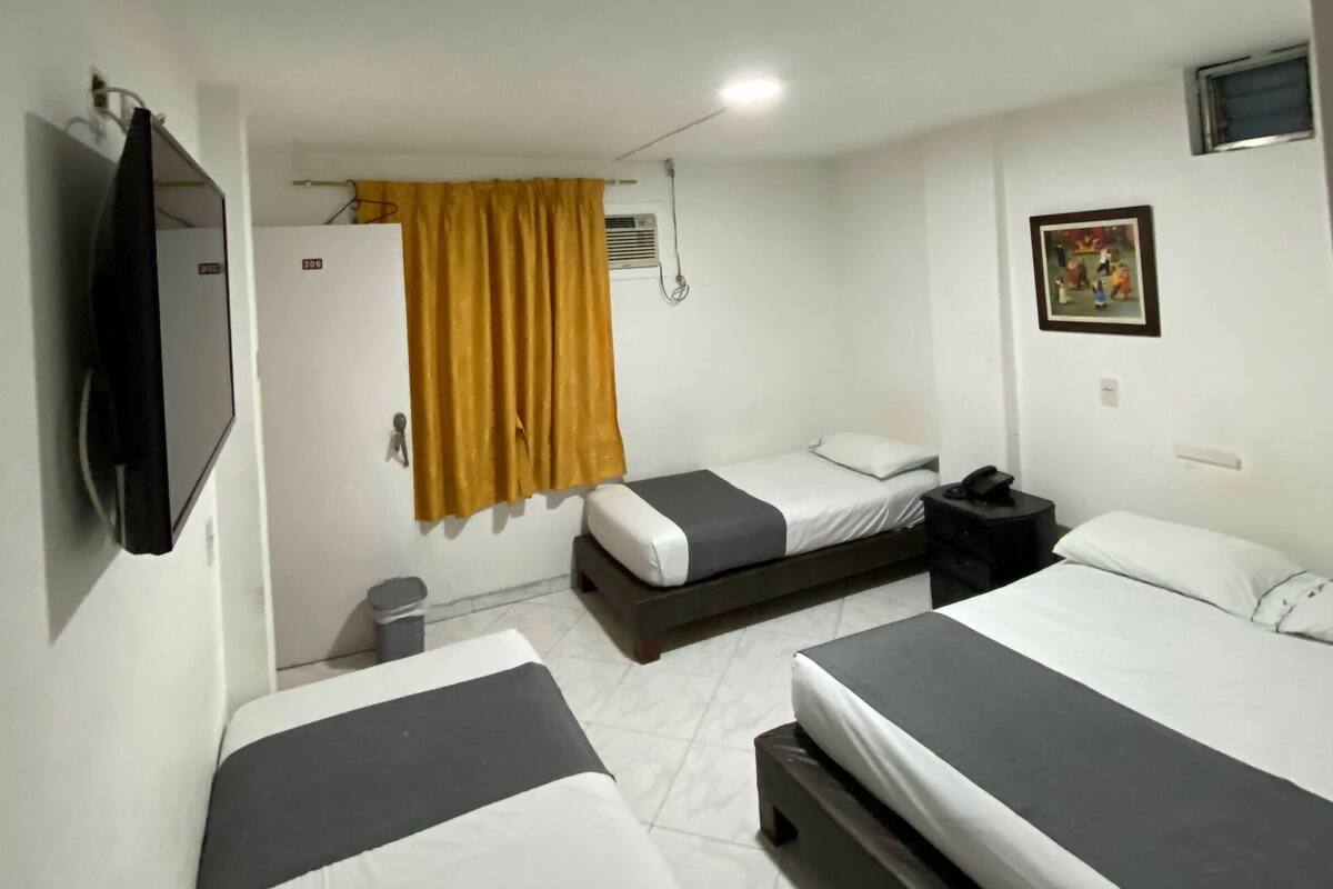 A hotel room with two single beds, a TV mounted on the wall, a small desk with a chair, and a window with curtains.