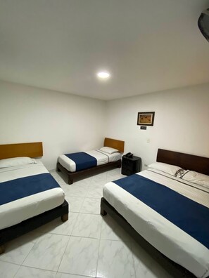 A hotel room with three beds, each with a wooden headboard and white bedding with blue accents. There is a small bedside table with a lamp and a phone on it. The room has white walls and a tiled floor. - HOTEL GRAN IMPERIO (Medellín)